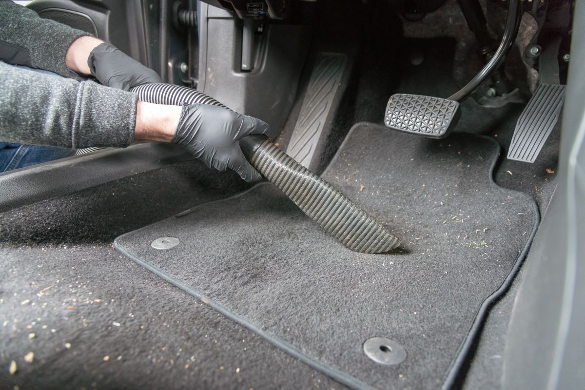 Gloved hands vacuuming the floor of a car, with a few food crumbs visible